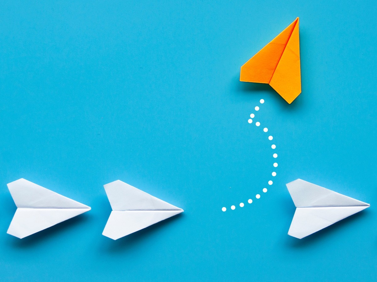 weiße Papierflieger in Reihe, ein orangener wechselt die Richtung